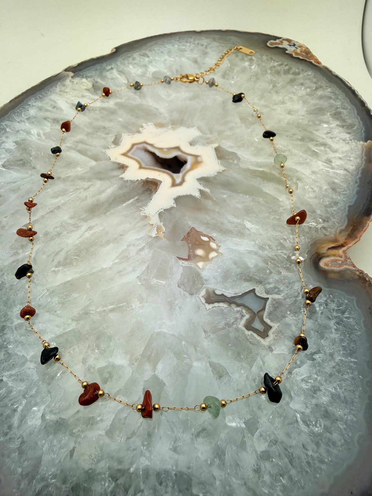 Handmade gold chain necklace resting on a polished white crystalline agate geode slice featuring irregular gemstone chips in jet black, terracotta, and shimmering goldstone with delicate gold bead accents