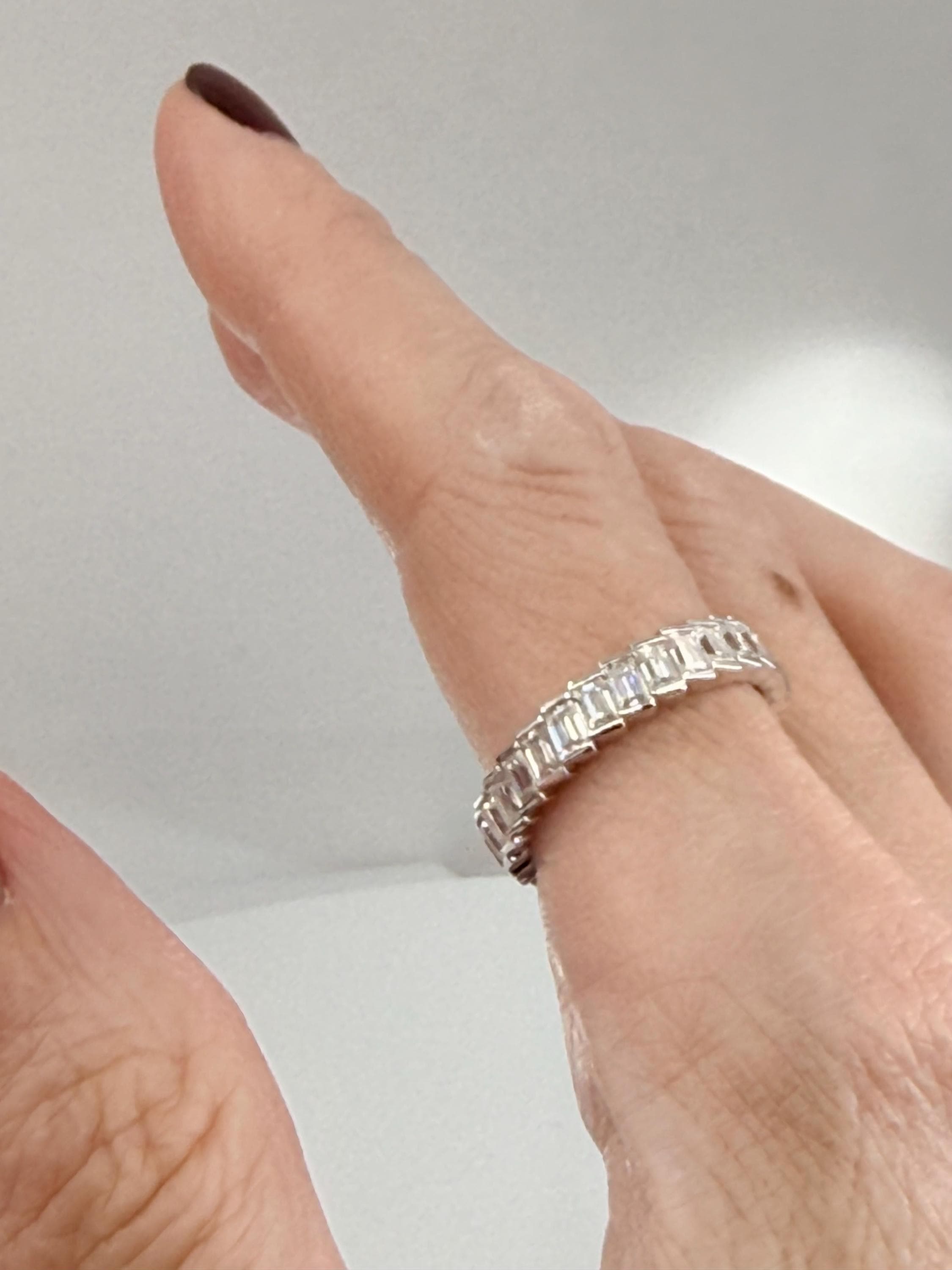 Digital photograph of a sparkling emerald-cut diamond eternity band worn on a hand with dark plum nail polish featuring clear step-cut stones set in white gold against a soft gray backdrop
