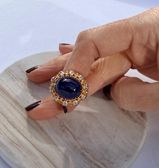 Fine jewelry photograph of a large deep blue oval cabochon sapphire ring with a gold filigree halo on a hand with dark burgundy nail polish resting atop a white veined marble surface