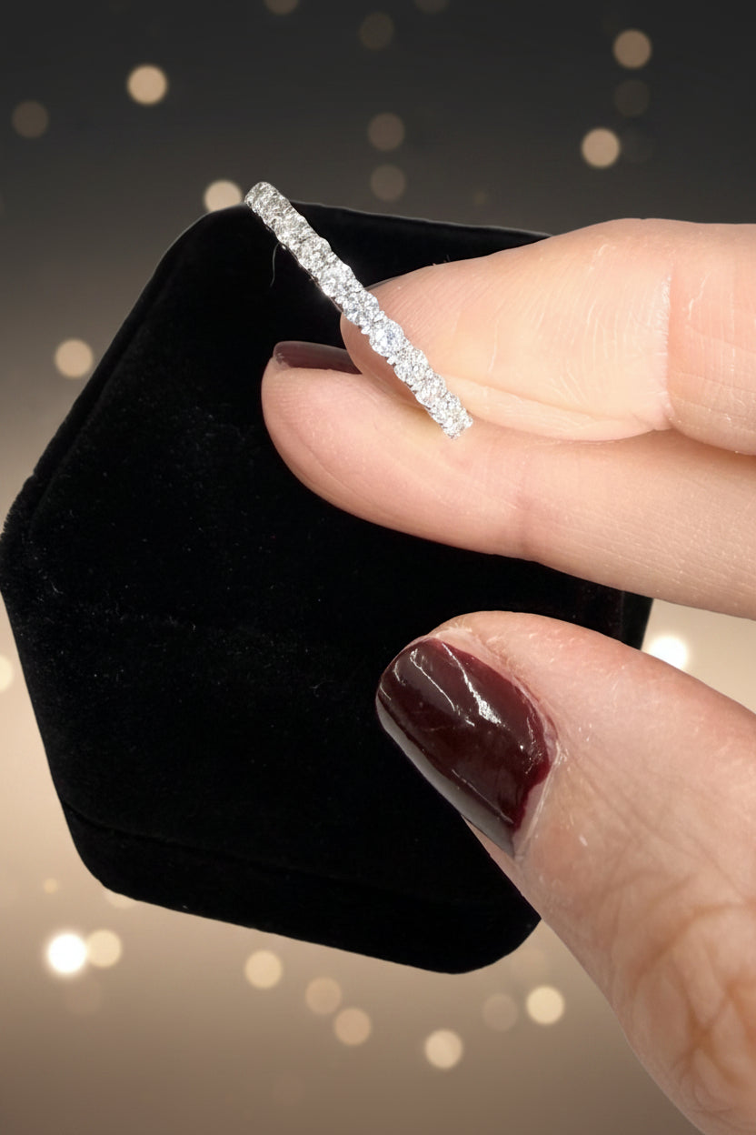 Photograph of a hand with deep burgundy nail polish holding a sparkling diamond eternity band against a black velvet box and a backdrop of warm golden bokeh lights