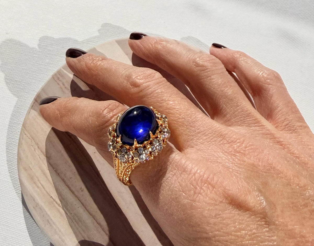 Gold and gemstone ring featuring a large deep blue cabochon sapphire and intricate filigree band worn on a hand with dark burgundy nails resting on a textured stone plate