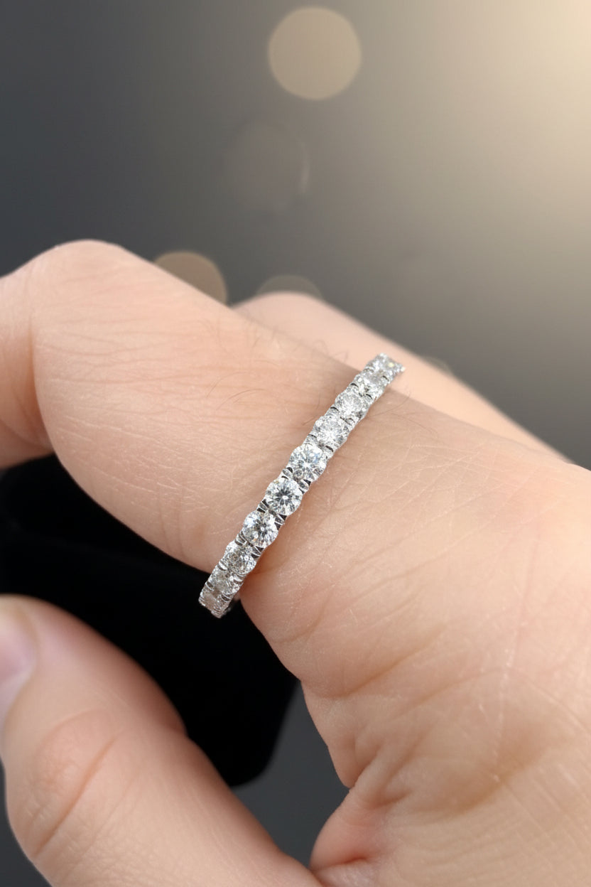 Photograph of a diamond eternity band worn on a finger featuring round brilliant-cut stones in a white gold prong setting against a blurred dark background with golden bokeh highlights