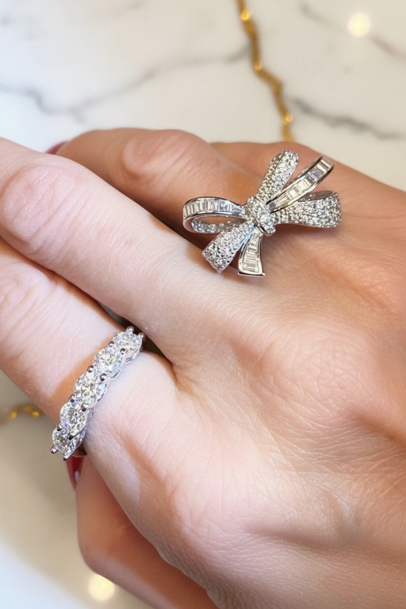 Photograph of a hand displaying an intricate diamond bow ring with baguette-cut stones and a shimmering eternity band against a white marble background with a gold chain